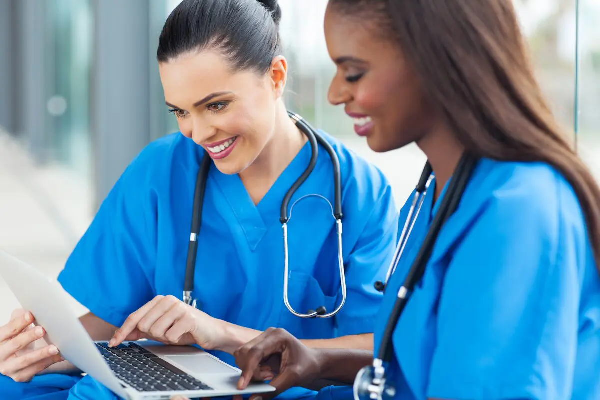 Two women working on a laptop and smiling wearing doctor's scrubs and stethoscopes