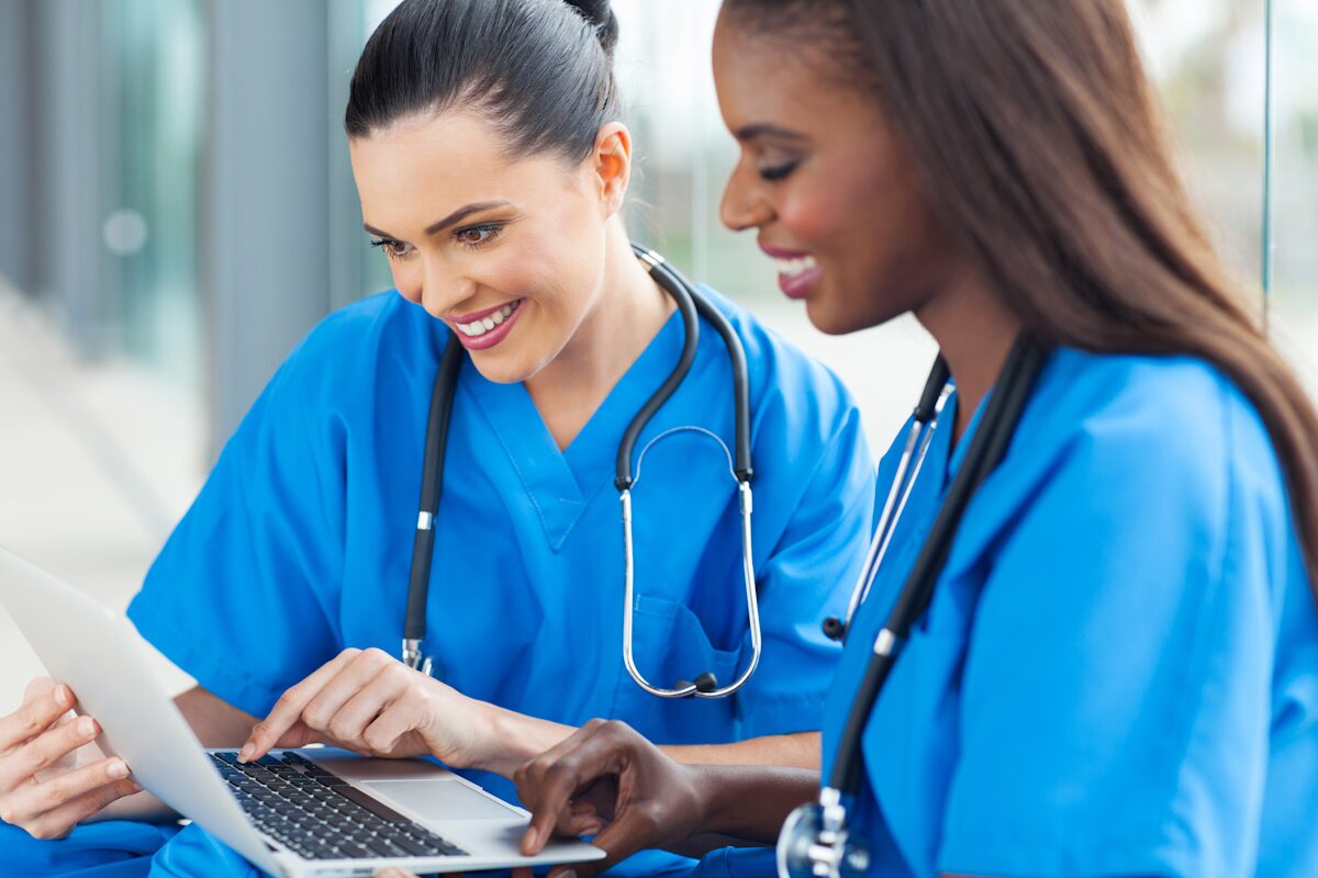 Two women working on a laptop and smiling wearing doctor's scrubs and stethoscopes